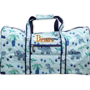 Pu&ograve; includere: Una borsa da viaggio azzurra con un motivo a dinosauri, con varie illustrazioni di dinosauri, palme e impronte. Il nome "Dean" &egrave; ricamato in arancione. La borsa ha manici e finiture blu navy.