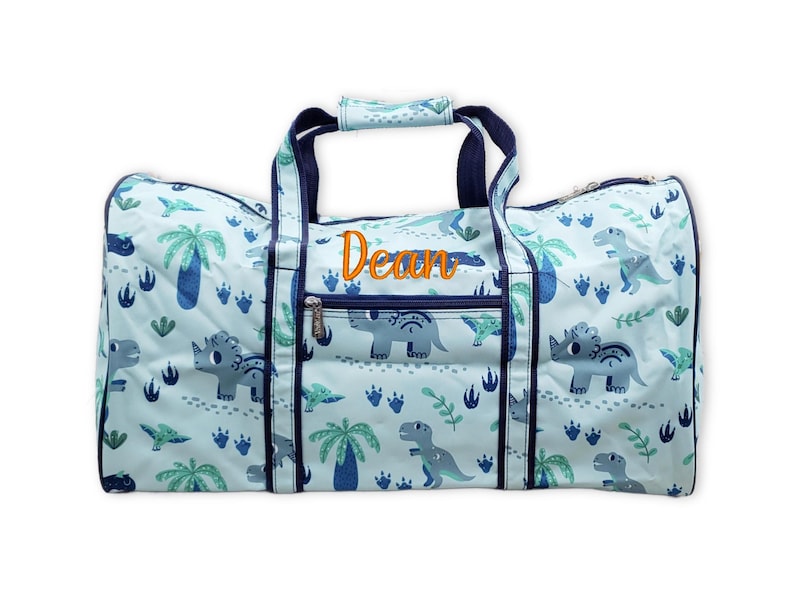 Pu&ograve; includere: Una borsa da viaggio azzurra con un motivo a dinosauri, con varie illustrazioni di dinosauri, palme e impronte. Il nome "Dean" &egrave; ricamato in arancione. La borsa ha manici e finiture blu navy.
