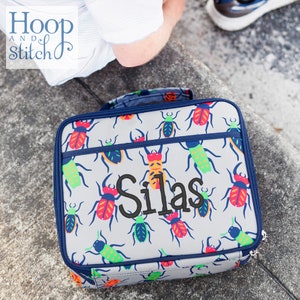 May include: A grey lunch bag with a blue trim and a zip closure. The bag has a repeating pattern of colourful bugs and the name "Silas" embroidered in black on the front.