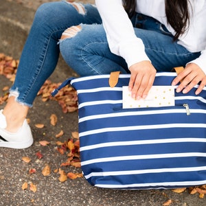 May include: A navy blue and white striped tote bag with a zipper closure and tan leather handles. A person is reaching into the front pocket of the bag to retrieve a small card. The bag is sitting on a concrete surface.