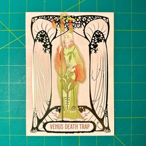 May include: A vintage-style illustration of a person standing in front of a Venus flytrap plant. The illustration is in a gold frame with a pink background. The text "VENUS DEATH TRAP" is at the bottom of the image.