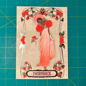 May include: A vintage-style illustration of a woman wearing a pink dress with red roses around her neck and holding a single red rose. The illustration is framed with a black and gold border with red roses and thorns. The text "THORNBACK" is printed in a pink banner at the bottom of the image.
