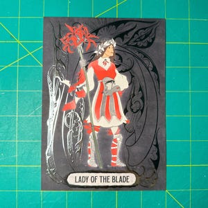 May include: A black and silver card with a woman in a red and white dress holding a staff with red flowers. The card has the text "Lady of the Blade" on it.