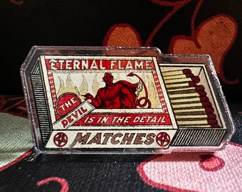 Puede incluir: Una caja de cerillas roja y negra con una cubierta de plástico transparente. La caja de cerillas tiene una ilustración de un diablo rojo con el texto "The Devil is in the Detail" y "Eternal Flame Matches".