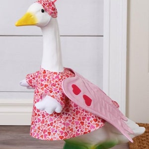 May include: A decorative goose figurine, painted white with a yellow beak, wearing a pink heart-patterned dress and headpiece. The goose has pink wings with red heart accents and stands on a green base, perfect for Valentine's Day decor.