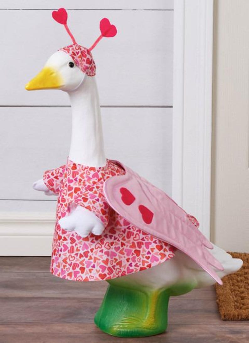 May include: A decorative goose figurine, painted white with a yellow beak, wearing a pink heart-patterned dress and headpiece. The goose has pink wings with red heart accents and stands on a green base, perfect for Valentine's Day decor.