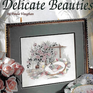 May include: A framed cross-stitch pattern book titled "Delicate Beauties" by Paula Vaughan. The image features a framed cross-stitch of a floral arrangement and a needlepoint hoop. The book cover includes the Leisure Arts logo and the text "Leaflet 2742 Book Fifty-Nine".