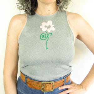 May include: A gray tank top with a floral design, paired with high-waisted blue jeans and a brown belt. The belt has a large, square buckle. The person is wearing sunglasses.
