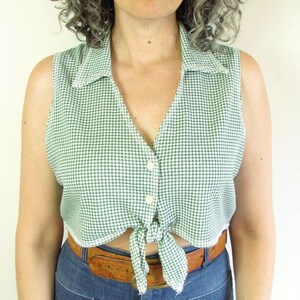 May include: A woman wearing a green and white gingham sleeveless top tied at the waist, paired with high-waisted blue jeans and a brown belt. She is also wearing large, round sunglasses.