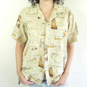 May include: Beige button-down shirt with a lighthouse and map print. The short-sleeved shirt features a collared neckline and a pattern of brown and white illustrations. The shirt is worn with denim shorts.