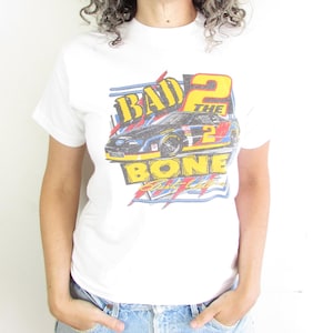 May include: White t-shirt with a graphic of a race car and the text "BAD 2 THE BONE". The shirt has a crew neck and short sleeves. The person is wearing sunglasses and denim shorts.