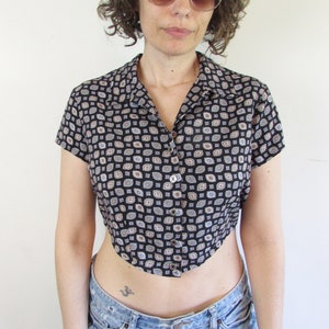 May include: A woman wearing a black and brown patterned cropped shirt with short sleeves and a button closure. She is also wearing light blue denim shorts.