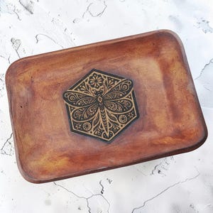 May include: A rectangular wooden tray with a smooth, warm brown finish. Centered on the tray is a black hexagonal inlay featuring a detailed, intricate laser-engraved design of a dragonfly surrounded by floral and geometric patterns.