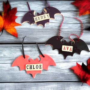 May include: Three ceramic bat-shaped ornaments in purple, black, and coral. Each bat has a name: "HENRY", "AVA", and "CHLOE" on a rectangular label. The ornaments have a glossy finish and are suspended by ribbons. Autumn leaves are scattered around.