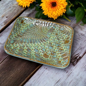 May include: Rectangular ceramic dish with a detailed lace pattern in shades of turquoise and brown. The edges are outlined in gold. The dish is displayed on a rustic wooden surface, with yellow flowers in the background.