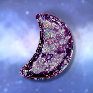 May include: A crescent-shaped ceramic dish with a glossy finish. The dish is primarily deep purple with speckled patterns of blue, green, and yellow. The rim is a darker shade of purple. The background is a gradient of blue and white.