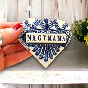 May include: A heart-shaped ceramic ornament featuring a white base with a dark blue lace-like pattern. A rectangular plaque is centered on the heart with the word NAGYMAMA embossed in dark blue capital letters. A dark blue ribbon loop is attached.