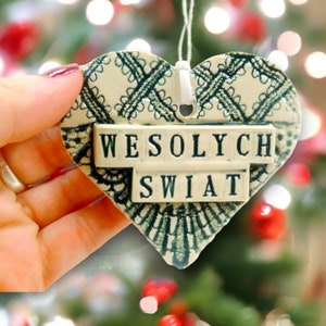Puede incluir: Un adorno de cer&aacute;mica blanca en forma de coraz&oacute;n con un dise&ntilde;o de encaje verde y las palabras "Wesołych Świąt" en letras negras.