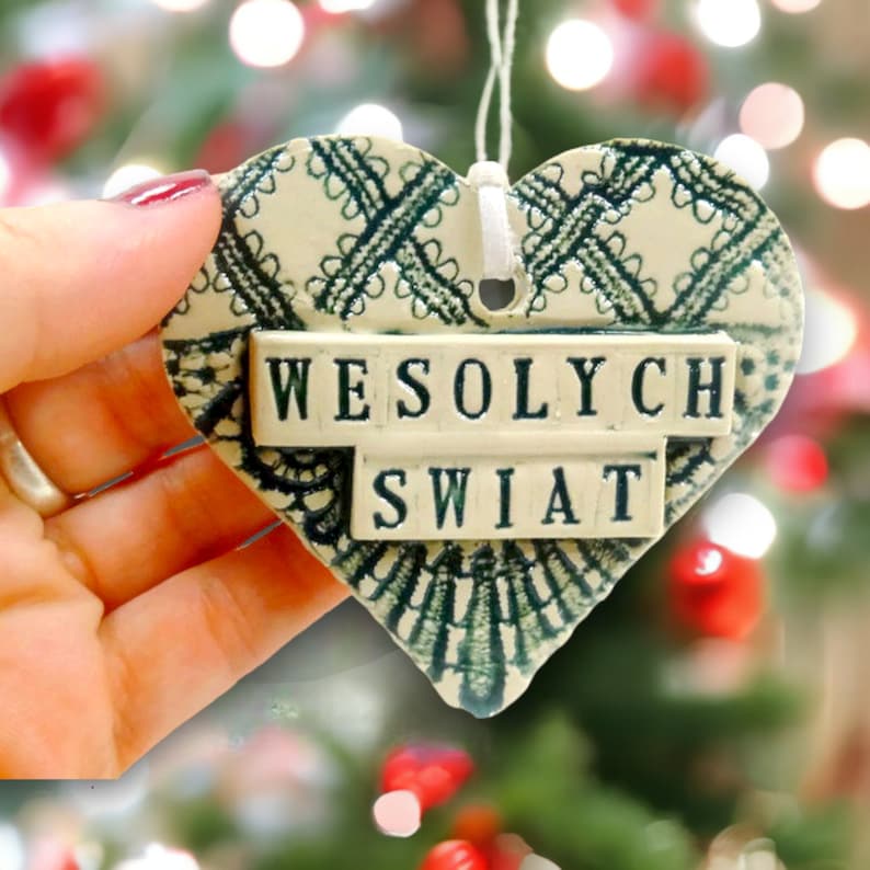 Puede incluir: Un adorno de cer&aacute;mica blanca en forma de coraz&oacute;n con un dise&ntilde;o de encaje verde y las palabras "Wesołych Świąt" en letras negras.
