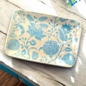 May include: Rectangular ceramic serving tray with a cream-colored background and a blue floral pattern. The tray features a repeating design of stylized flowers, leaves, and berries. The tray is placed on a white wooden surface.