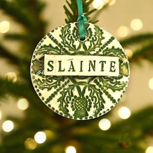 May include: A white ceramic Christmas ornament with a green Celtic knot design and the word "Slainte" in white lettering. The ornament is hanging from a green ribbon on a Christmas tree.