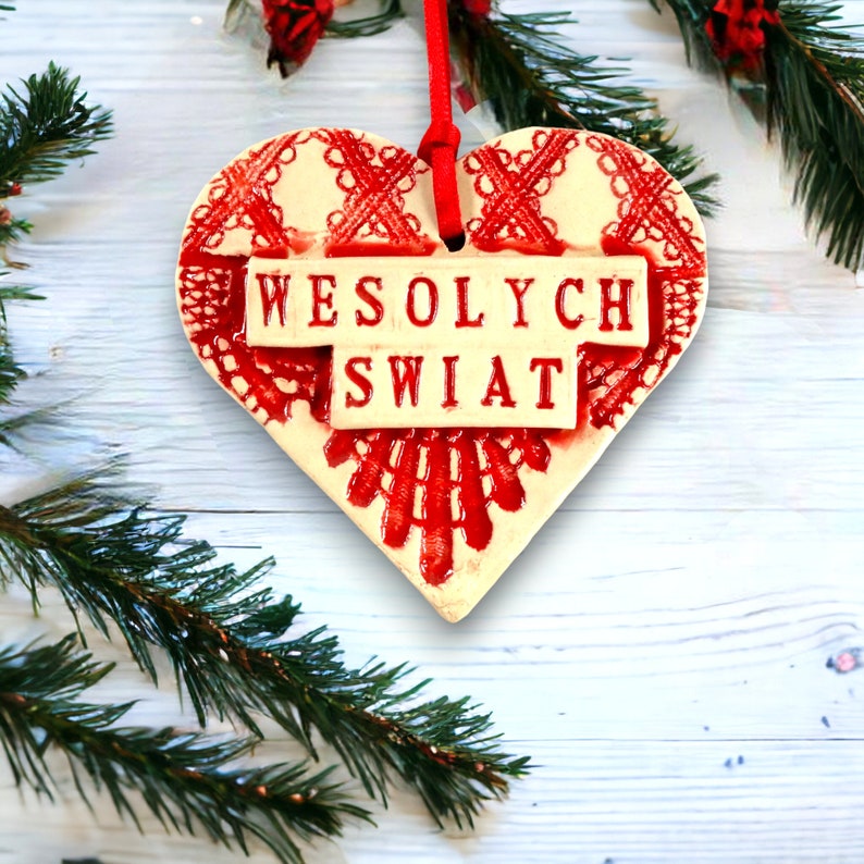 Puede incluir: Un adorno de cer&aacute;mica blanca en forma de coraz&oacute;n con detalles de encaje rojo y las palabras "Wesołych Świąt" en letras rojas.