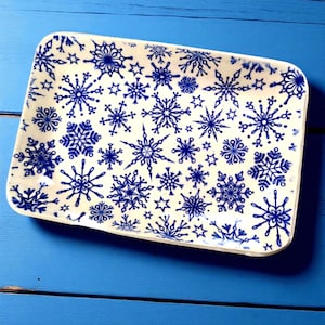 Puede incluir: Una fuente rectangular con base crema, adornada con un patrón repetitivo de copos de nieve y estrellas azules. La fuente tiene esquinas redondeadas y está sobre un fondo azul, sugiriendo un tema invernal o festivo.