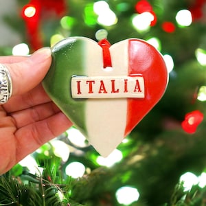 Puede incluir: Un adorno de cerámica en forma de corazón con los colores verde, blanco y rojo de la bandera italiana. Una placa rectangular blanca está fijada en el centro con la palabra ITALIA impresa en letras rojas. Una cinta roja está en la parte superior.