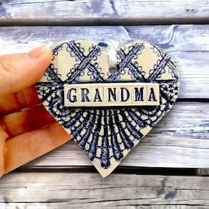 Puede incluir: Adorno de cerámica en forma de corazón con un diseño de encaje azul y blanco. La palabra "GRANDMA" se muestra en letras azules en bloque sobre una placa rectangular blanca. Se adjunta una cinta blanca para colgar.