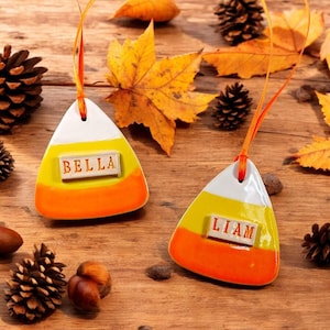 May include: Two ceramic candy corn ornaments with the names "BELLA" and "LIAM" on them. The ornaments are white, yellow, and orange with orange ribbons for hanging. The background includes fall leaves, pinecones, and nuts.