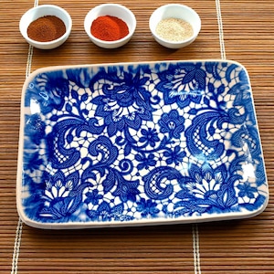 May include: A rectangular ceramic serving tray with a blue and white floral lace design. Three small white bowls hold spices: brown, red, and white. The tray is on a bamboo mat.