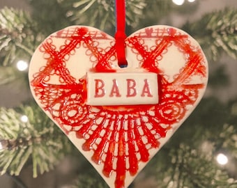 Könnte beinhalten: Ein herzförmiges Keramikornament mit einem roten spitzenartigen Muster auf weißem Untergrund. In der Mitte befindet sich eine kleine rechteckige Plakette mit dem in Rot gedruckten Wort BABA. Ein rotes Band ist oben zur Aufhängung befestigt.