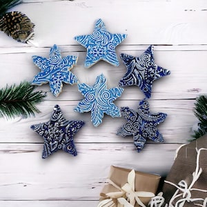 Puede incluir: Seis adornos azules en forma de estrella con diseños de copos de nieve y remolinos blancos. Los adornos están dispuestos sobre una superficie de madera blanca, con cajas de regalo y ramas de pino en el fondo. Ideales para la decoración navideña.
