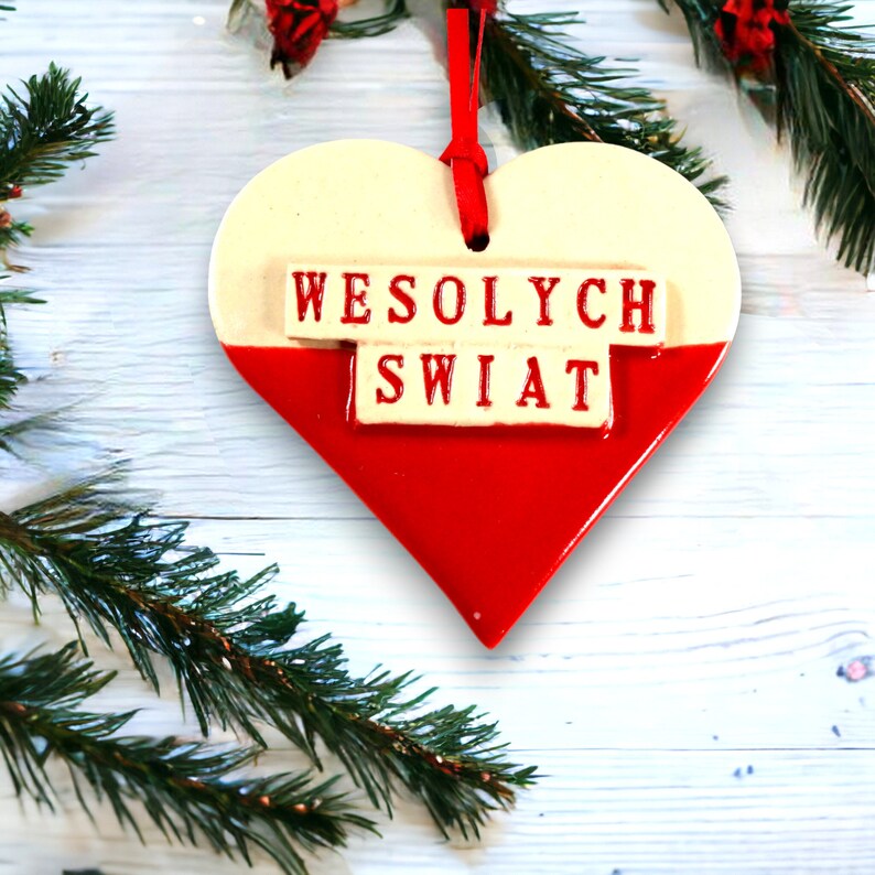 Puede incluir: Un adorno en forma de coraz&oacute;n blanco y rojo con la frase polaca "Wesołych Świąt" escrita en rojo. El adorno est&aacute; colgado de una cinta roja.