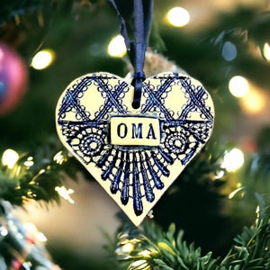 May include: Cream and blue heart-shaped ceramic Christmas ornament with a lace design. The word "OMA" is displayed on a rectangular plaque. A dark blue ribbon is attached for hanging. The ornament is on a Christmas tree with fairy lights.