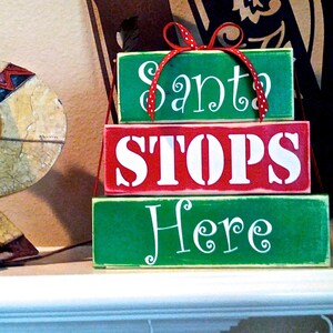 May include: Three wooden blocks stacked on top of each other. The top block is green and says "Santa" in white cursive writing. The middle block is red and says "STOPS" in white block letters. The bottom block is green and says "Here" in white cursive writing. A red ribbon with white polka dots is tied around the top block.