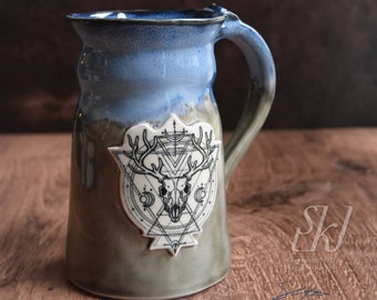 May include: A ceramic mug with a blue and green glaze. The mug features a white decal of a skull with antlers inside a geometric design. The mug is sitting on a wooden surface.