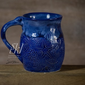 May include: A blue ceramic mug with a white glaze and a raised design of seashells and starfish. The mug has a rounded body and a handle.
