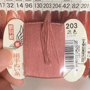 Può includere: Un'bobina di filo da ricamo rosa con un'etichetta che recita "203 誘色 (buiro)" e "絹手ぬい糸 (kinu tenui ito)". L'etichetta include anche un codice a barre e il testo "④大黑絲業株式会社 (Dai-koku Shigyo Kabushiki Kaisha)".