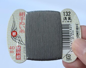May include: A spool of dark gray silk sewing thread wrapped around a flat, cream-colored cardboard card. The card features Japanese text, the number 132, a barcode, and a graphic of a wing. The thread is labeled as 40 meters and 100% silk.