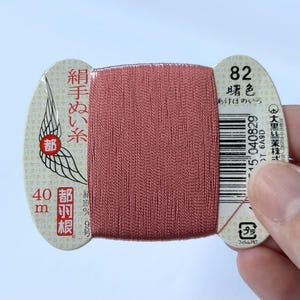 以下が含まれることがあります： クリーム色の厚紙に巻かれた、くすんだピンク色の縫い糸。厚紙には日本語のテキスト、数字の82、バーコード、および「40 m」という表記が印刷されています。糸は滑らかで、わずかに光沢のある質感をしています。