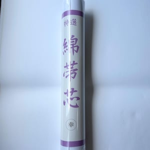 May include: A cylindrical roll of white fabric wrapped in a paper sleeve. The sleeve features a purple horizontal band at the top and bottom, with Japanese text printed in purple vertically down the center and a small circular logo at the bottom.