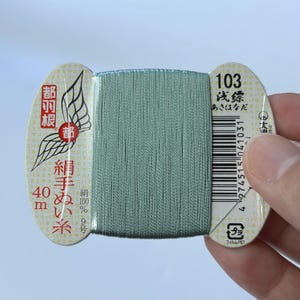 May include: A spool of light sage green silk sewing thread wrapped around a cream-coloured cardboard bobbin. The bobbin features black Japanese text, a wing graphic, the number 103, a barcode, and the text 40m, indicating the thread length.