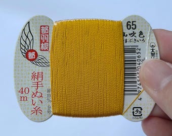 May include: A spool of vibrant golden yellow silk thread wrapped around a white cardboard card. The card features Japanese text, a wing logo, the number 65, and 40m printed on the left side. A barcode is visible on the right side of the card.