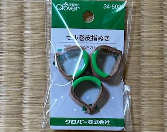 Op de afbeelding: Drie bruine lederen vingerhoeden met felgroene plastic randen, verpakt in een doorzichtige plastic hoes op een wit-groene kaart met het label Clover 34-503. De verpakking bevat Japanse tekst en een ophanggat aan de bovenzijde.