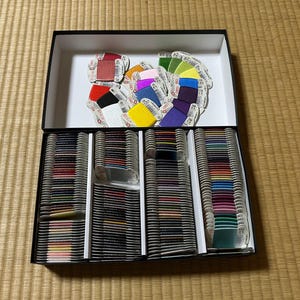 Könnte beinhalten: Eine große schwarze Aufbewahrungsbox mit einer umfangreichen Sammlung von Stickgarnspulen. Der untere Bereich ist in vier Fächer unterteilt, die mit ordentlich gestapelten, mehrfarbigen Garnspulen gefüllt sind, während der Deckel eine lose Auswahl verschiedenfarbiger Garnspulen enthält.
