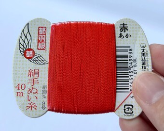 Op de afbeelding: Een klos felrode zijden draad gewikkeld rond een witte kartonnen kaart. De kaart bevat Japanse tekst, een vleugellogo, een streepjescode en de tekst 40m. De draad is 100% zijde, bedoeld voor handwerk, gepresenteerd op een platte, ovale kaart.