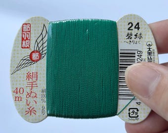 May include: A spool of vibrant emerald green silk thread wrapped around a beige cardboard card. The card features Japanese text, the number 24, a barcode, and the measurement 40 m. The thread is wound tightly in a vertical orientation.