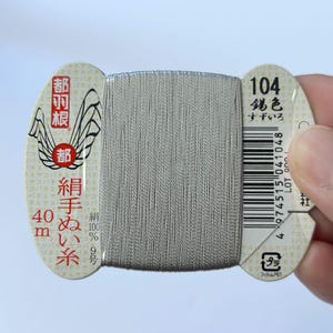 May include: A spool of silver-coloured thread wrapped around a branded cardboard card. The card features the text 104, 40m, 100% silk, and Japanese characters. The thread is wound vertically across the centre of the card, which has rounded edges.