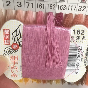 Könnte beinhalten: Eine rosa Stickgarnrolle mit der Nummer 162 und dem Text "菖蒲色 あやめいろ" (bedeutet "Iris Farbe") auf dem Etikett. Das Etikett enthält auch den Text "絹手ぬい糸" (bedeutet "Seidenhandnähgarn") und "都羽根" (bedeutet "Hauptfeder").